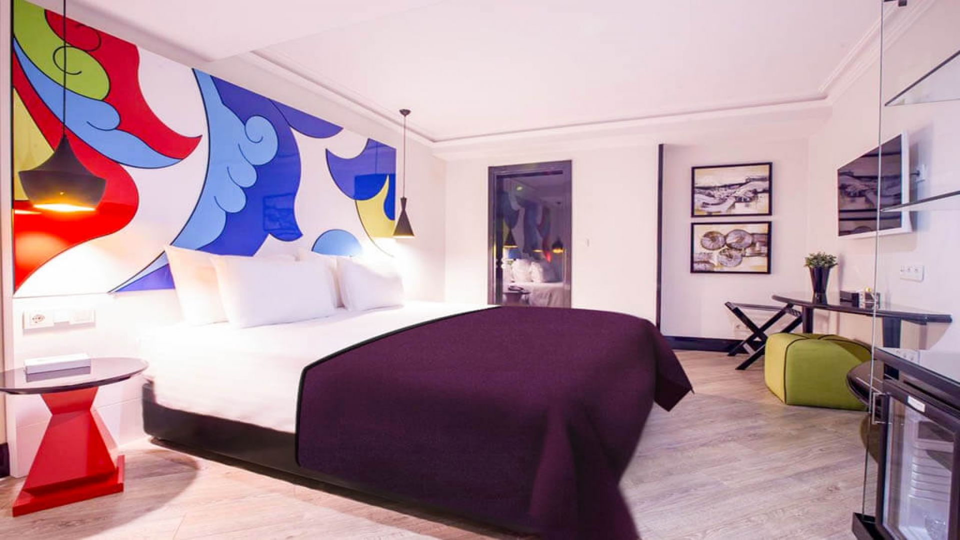 Modern hotel room with a vibrant, colorful mural above the bed, a purple bedspread, unique red side table, light wood flooring, and contemporary furnishings.