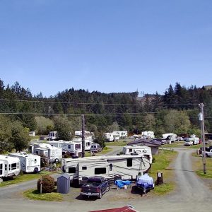 Aerial view of an RV park featuring numerous campers, vehicles including a yellow car, a central building, and gravel roads amidst grassy areas, with a forest in the background.