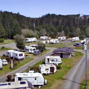 Aerial view of an RV park featuring numerous campers, vehicles including a yellow car, a central building, and gravel roads amidst grassy areas, with a forest in the background.