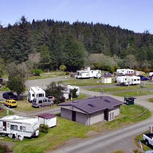 Aerial view of an RV park featuring numerous campers, vehicles including a yellow car, a central building, and gravel roads amidst grassy areas, with a forest in the background.