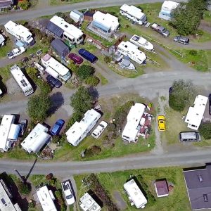 Aerial drone shot of a bustling RV park, showcasing many campers, motorhomes, and vehicles, including a distinctive yellow car, on gravel and grass lots.